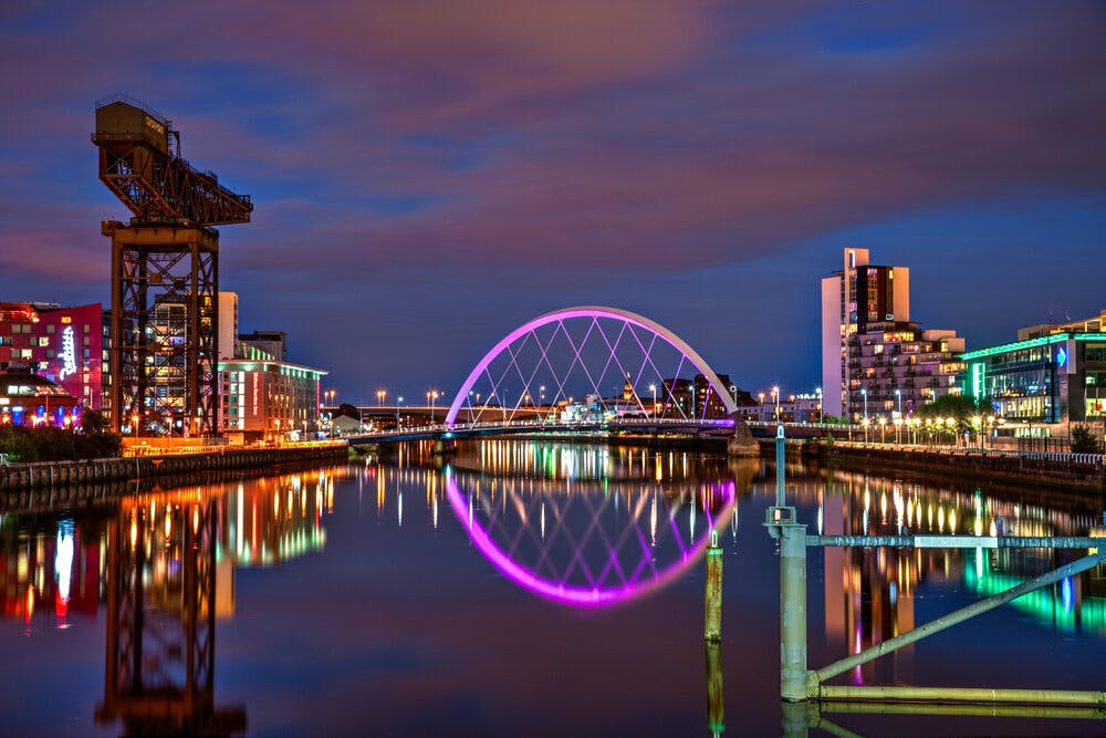 Twilight view of the Clyde Arc also known as the Squinty Bridge over the River Clyde in Glasgow with the city lights reflecting on the water and a historic crane on the left against a dusk sky