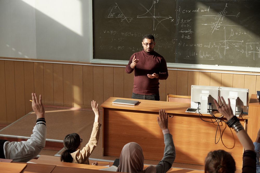 Mathematics lecture in progress with a male teacher explaining a concept,
students raising hands to ask questions, and complex equations written on the
blackboard in the
background.