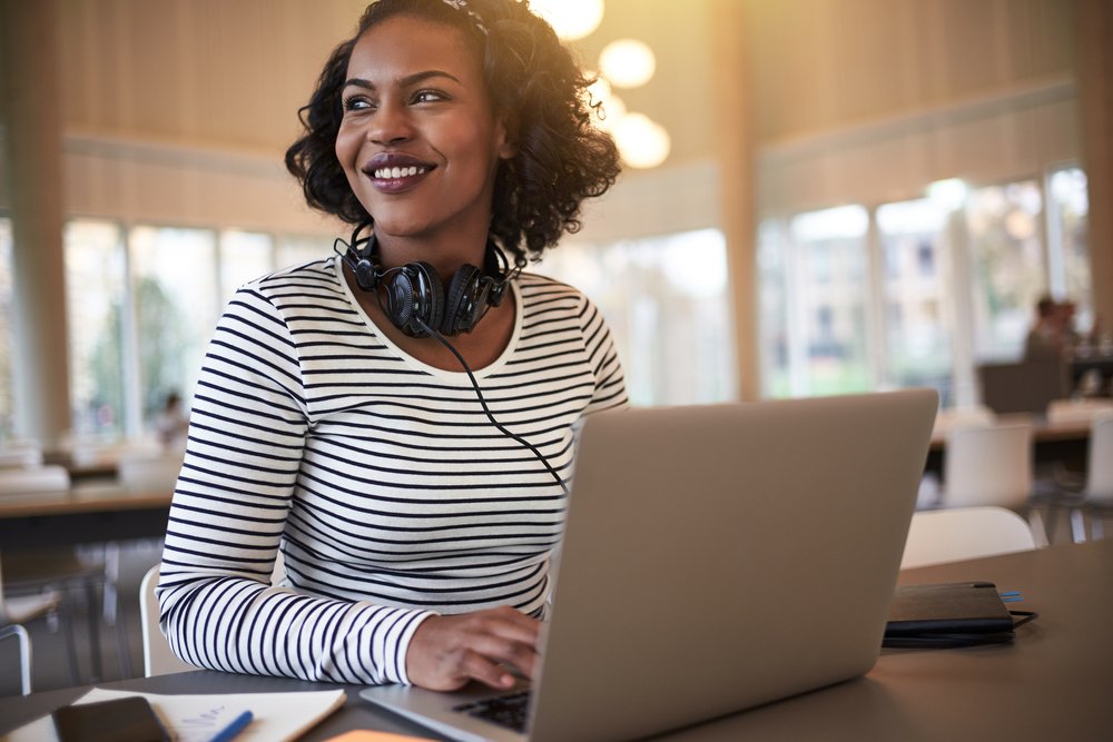 Smiling woman with headphones around her neck sitting at a table with a
laptop, notebook, and smartphone in a well-lit indoor space with large
windows